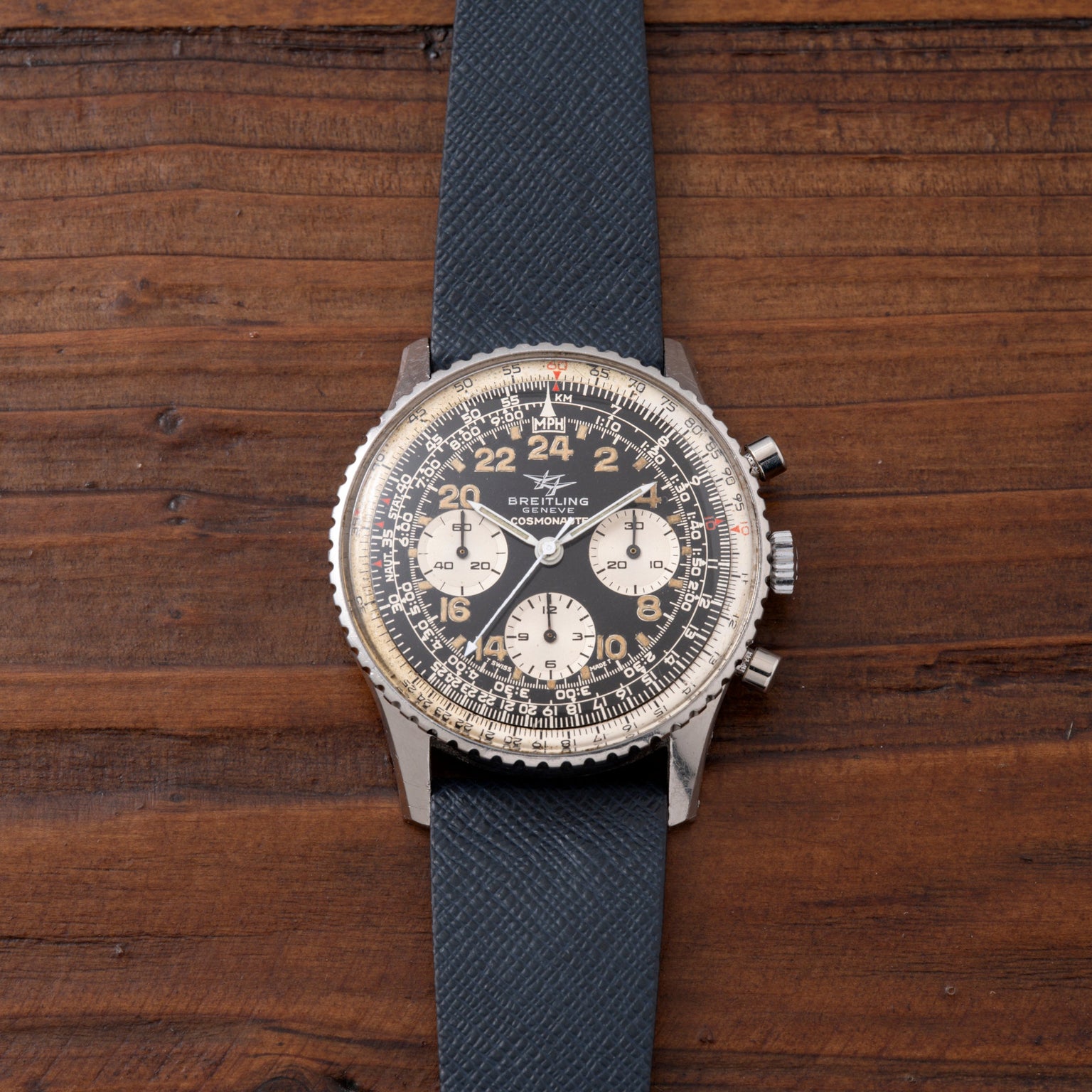 Breitling Cosmonaute chronograph watch with black dial, white subdials, and blue fabric strap on wooden surface.