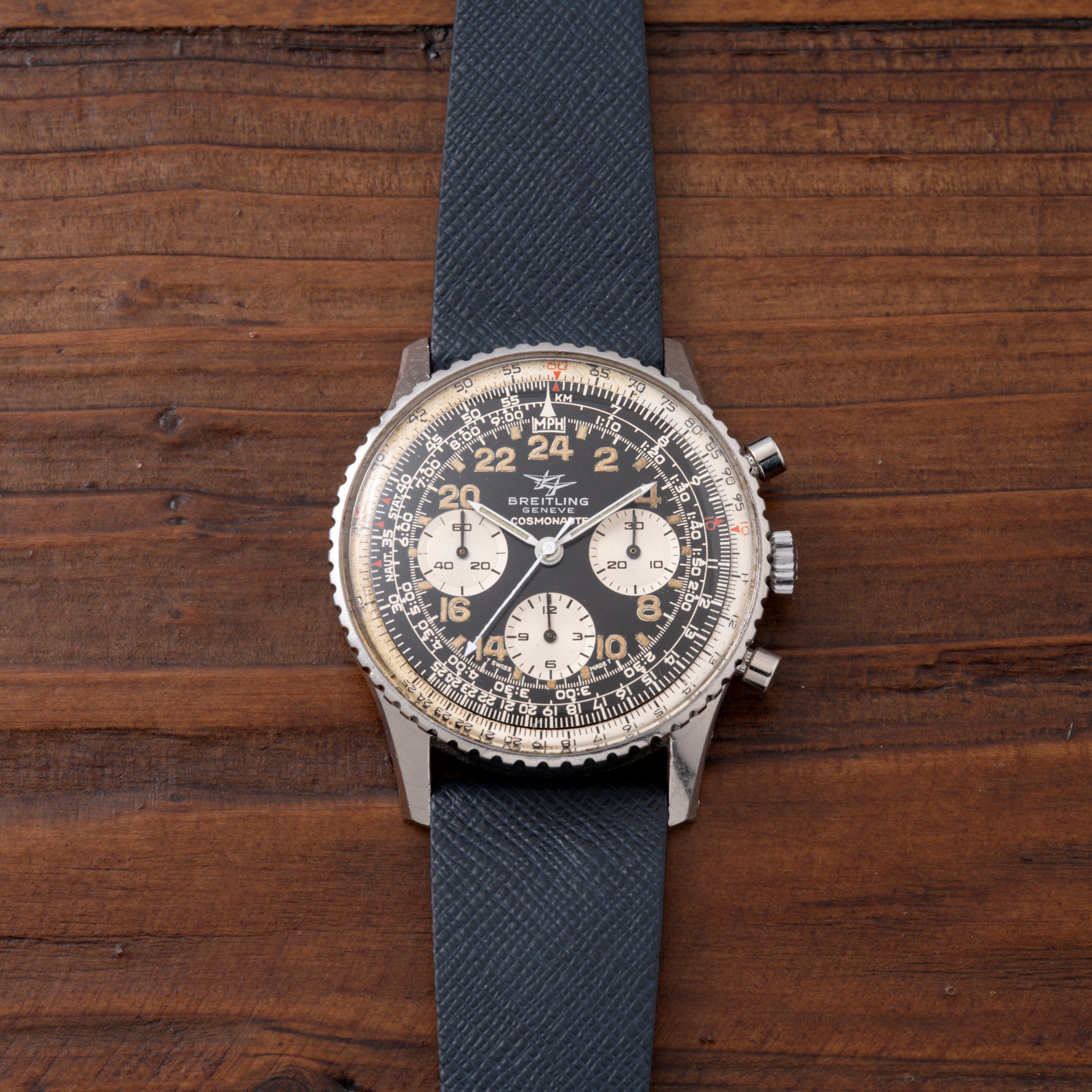 Breitling Cosmonaute chronograph watch with black dial, white subdials, and blue fabric strap on wooden surface.