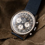Breitling Cosmonaute chronograph watch with black dial, gold numerals, two subdials, and blue fabric strap on neutral fabric