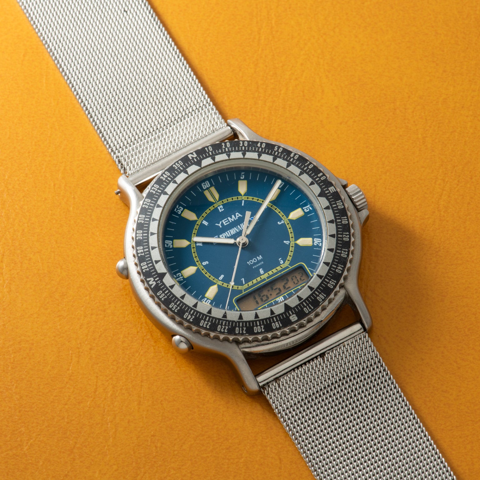 Yema Spationaute ISS watch with blue dial, luminous indices, rotating bezel, silver mesh strap, and digital subdial on yellow