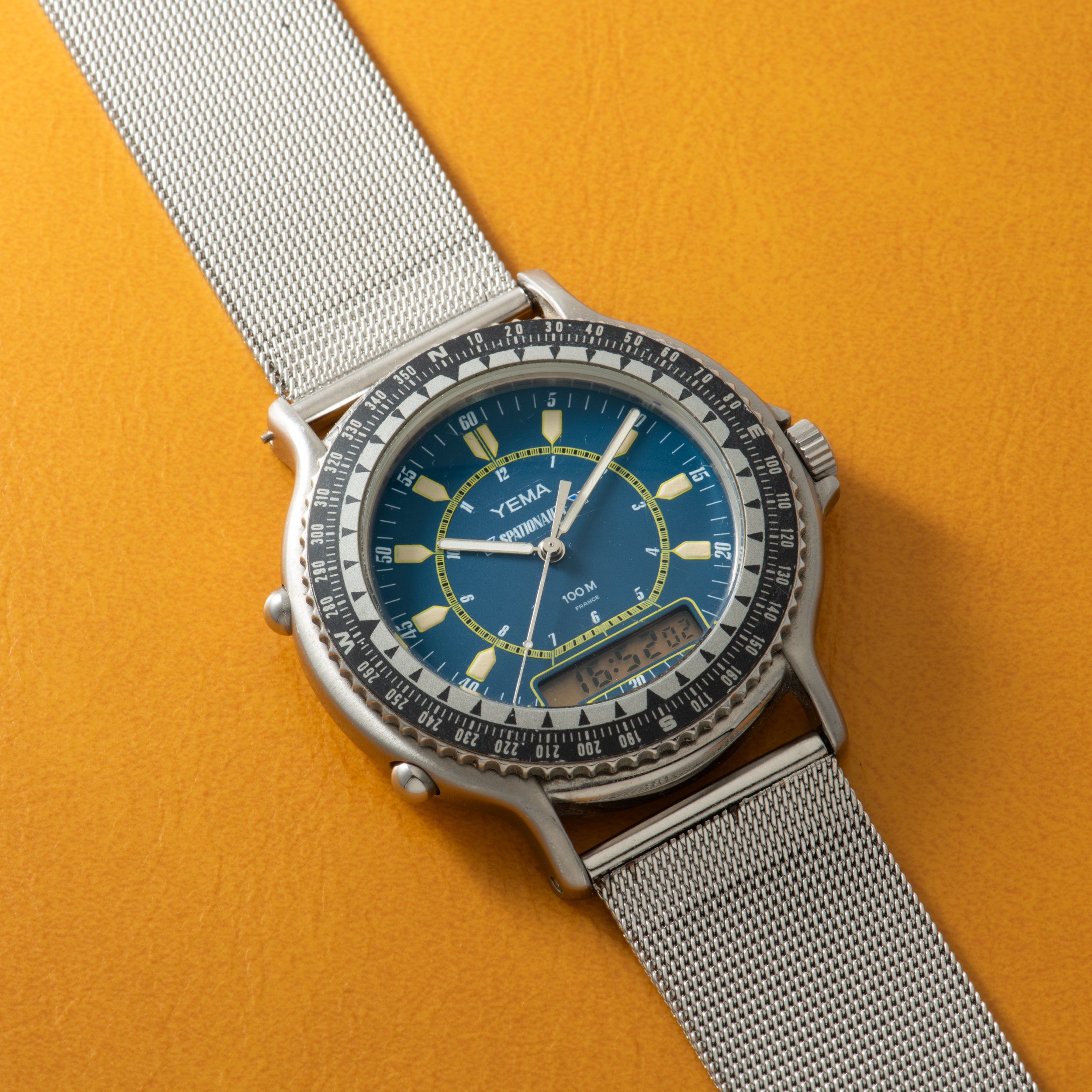 Yema Spationaute ISS watch with blue dial, luminous indices, rotating bezel, silver mesh strap, and digital subdial on yellow
