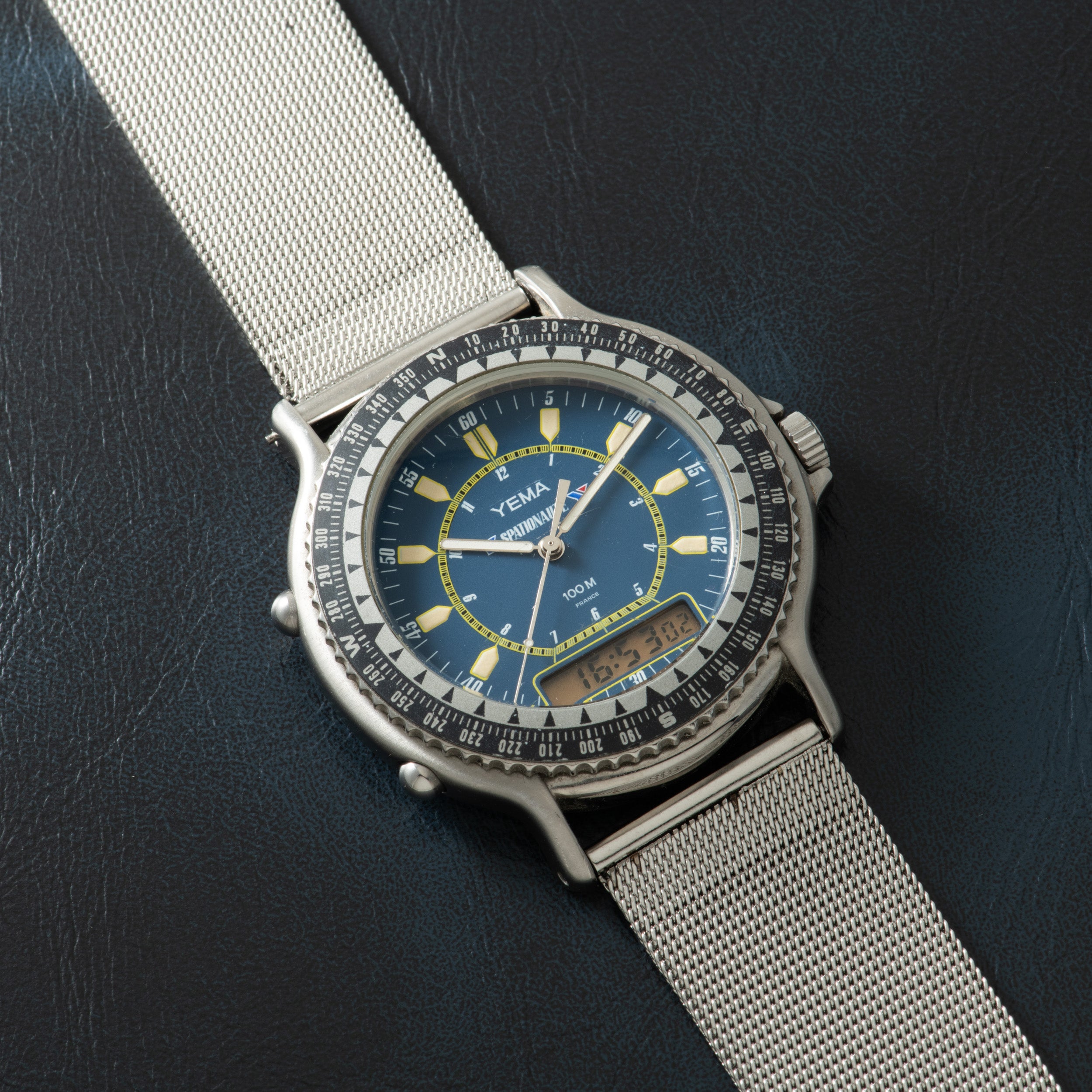 Silver-toned watch with blue dial, yellow hour markers, dual analog and digital displays, and white mesh band on dark surface