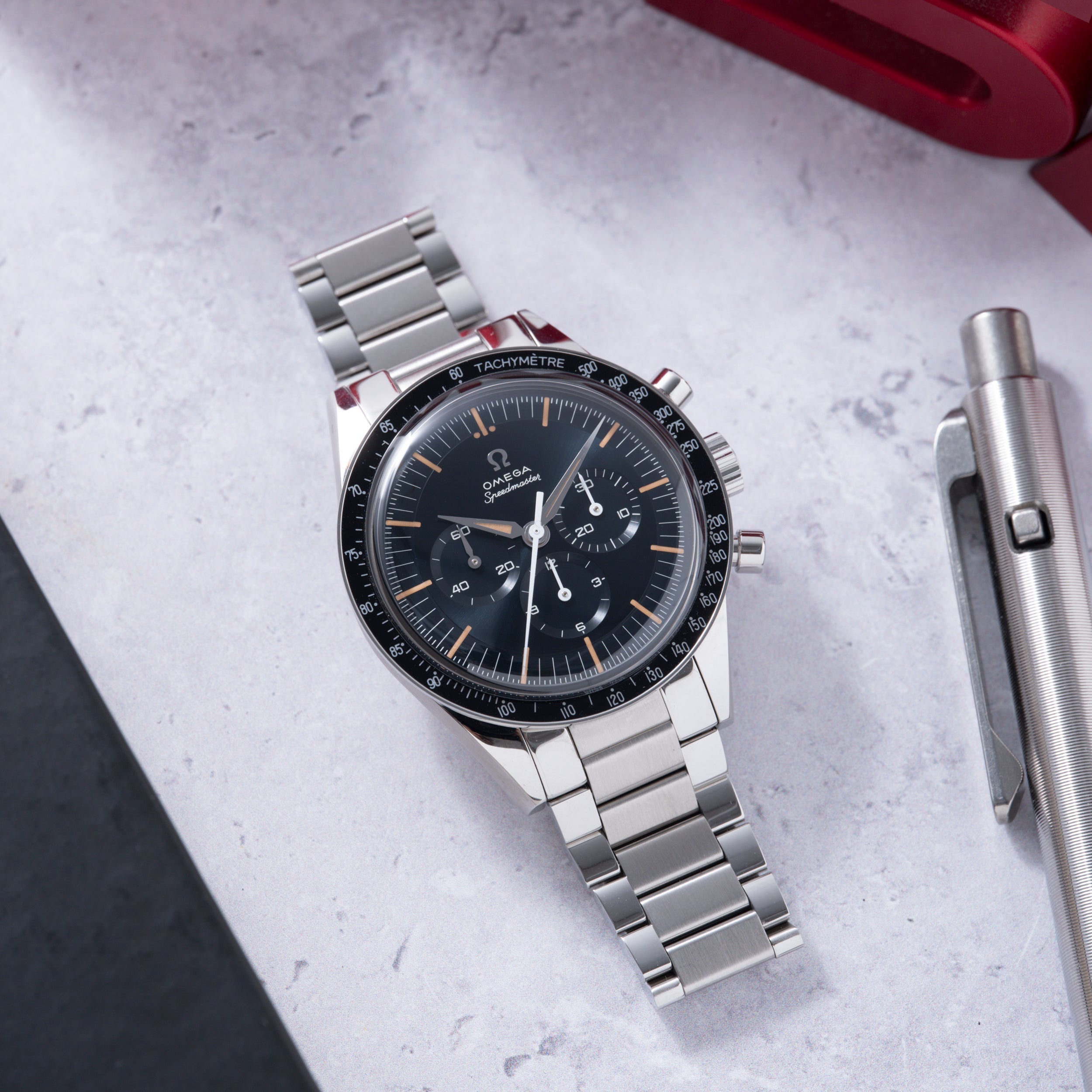 Omega Speedmaster chronograph watch with black dial, tachymetre bezel, steel bracelet, and orange hour markers on marble surf