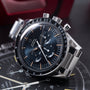 Stainless steel chronograph watch with black dial, tachymeter bezel, three subdials, and orange hour markers.