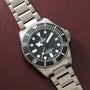 Steel dive watch with black dial, white hour markers, rotating bezel, and three-link bracelet on burgundy background.