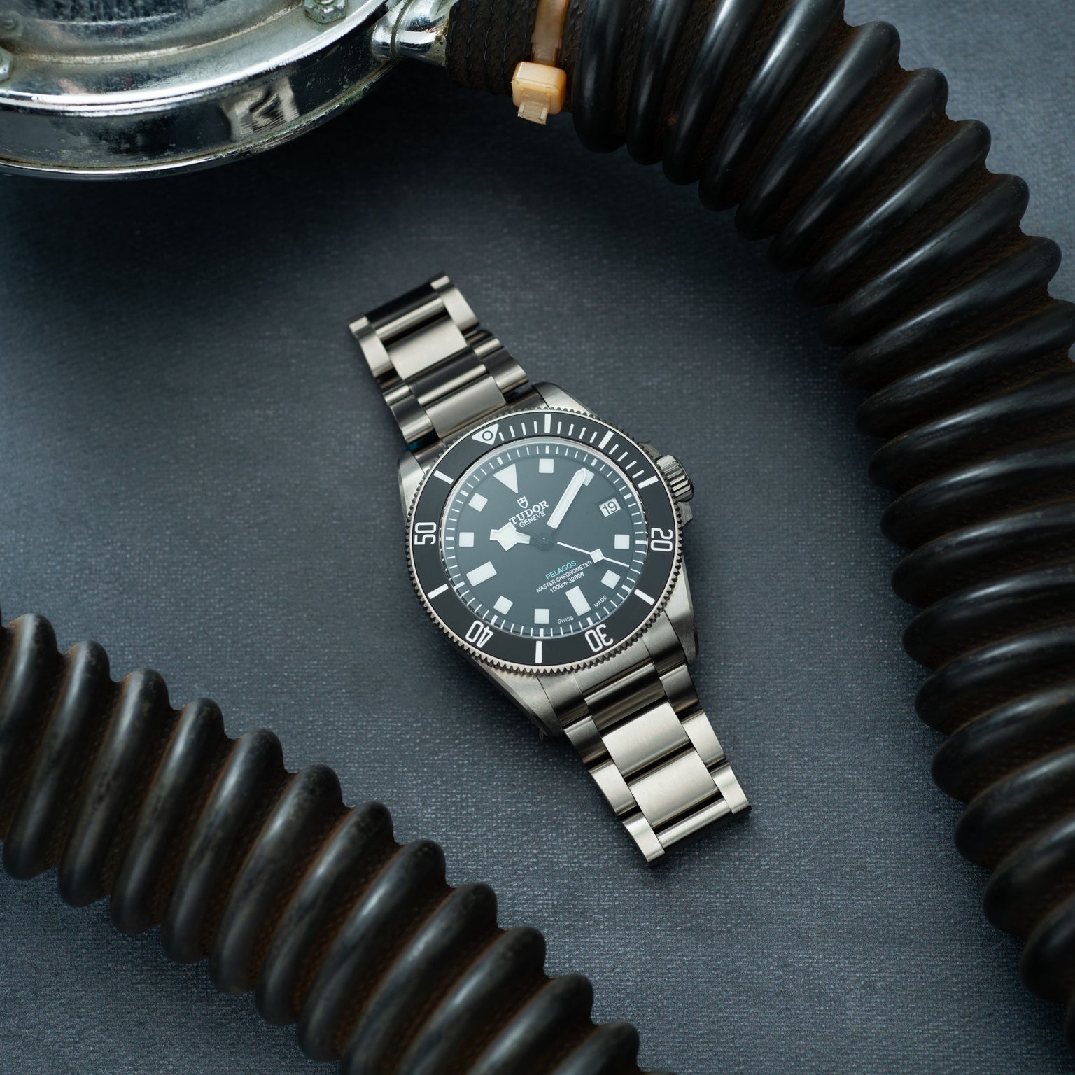 Tudor Pelagos Ultra dive watch with dark dial and stainless steel bracelet, displayed on blue surface surrounded by black coi