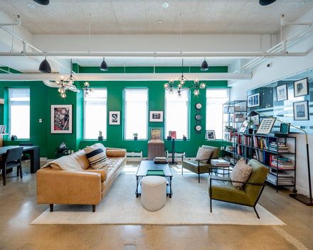 Modern lounge with green walls, brown sofa, two chairs, bookshelves, and large windows letting in light.