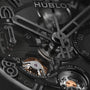 Black watch with visible Hublot branding, skeleton dial showing mechanical movement with gold accents, and skeletal hour and 