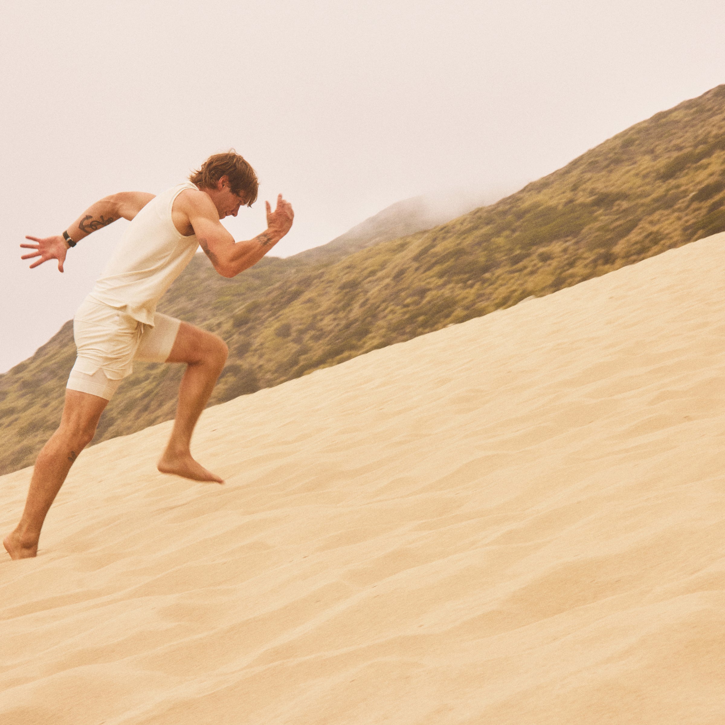 Man running barefoot up a sandy hill with hills and cloudy sky in the background.