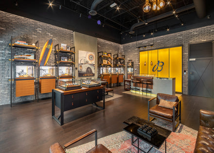 Modern watch boutique with leather chairs, display cases, gray brick walls, and warm yellow accent lighting.