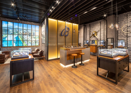 Modern watch boutique with display cases, stools at a central counter, and a large scenic mural on the wall.