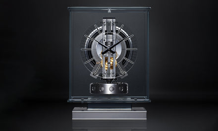 A modern, transparent glass clock with visible mechanical gears on a dark background.