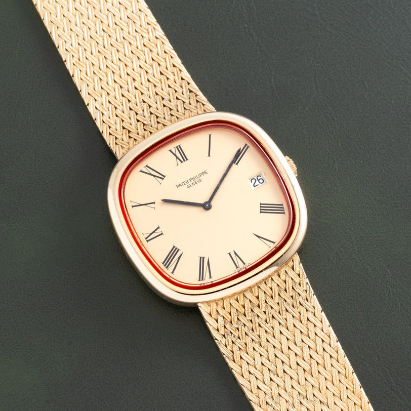 The Patek Philippe Ellipse TV Case watch, featuring Roman numerals and a date display on a woven gold band, rests on a dark surface.