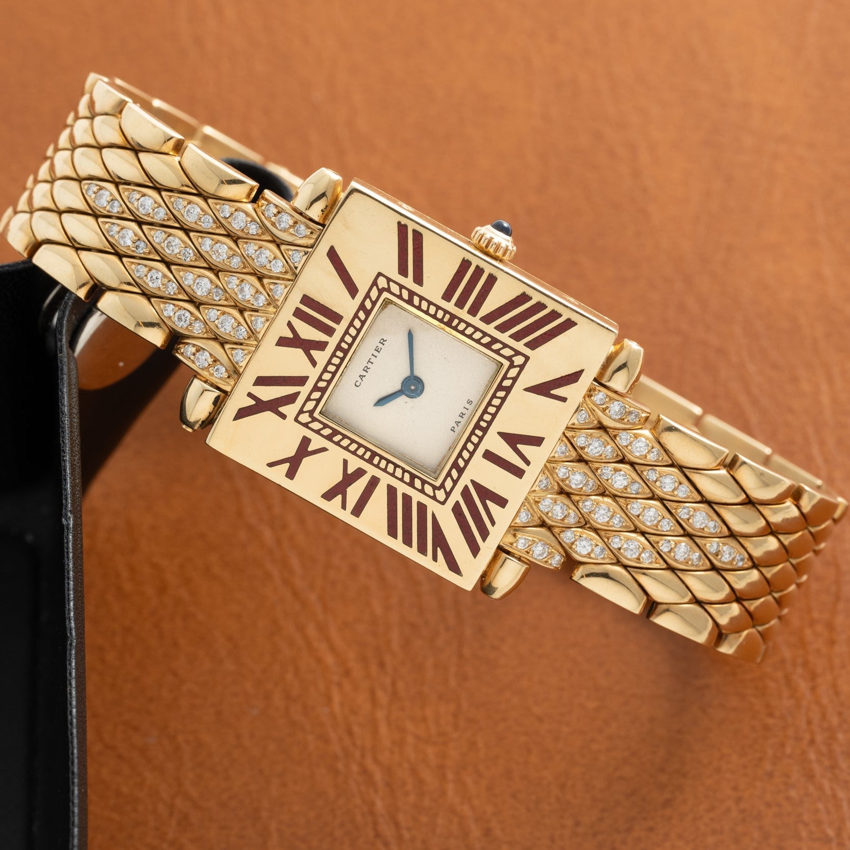Gold-tone watch with square cream dial, Roman numerals, diamond-set bracelet, and blue hands against tan background.