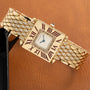 Gold-tone watch with square cream dial, Roman numerals, diamond-set bracelet, and blue hands against tan background.