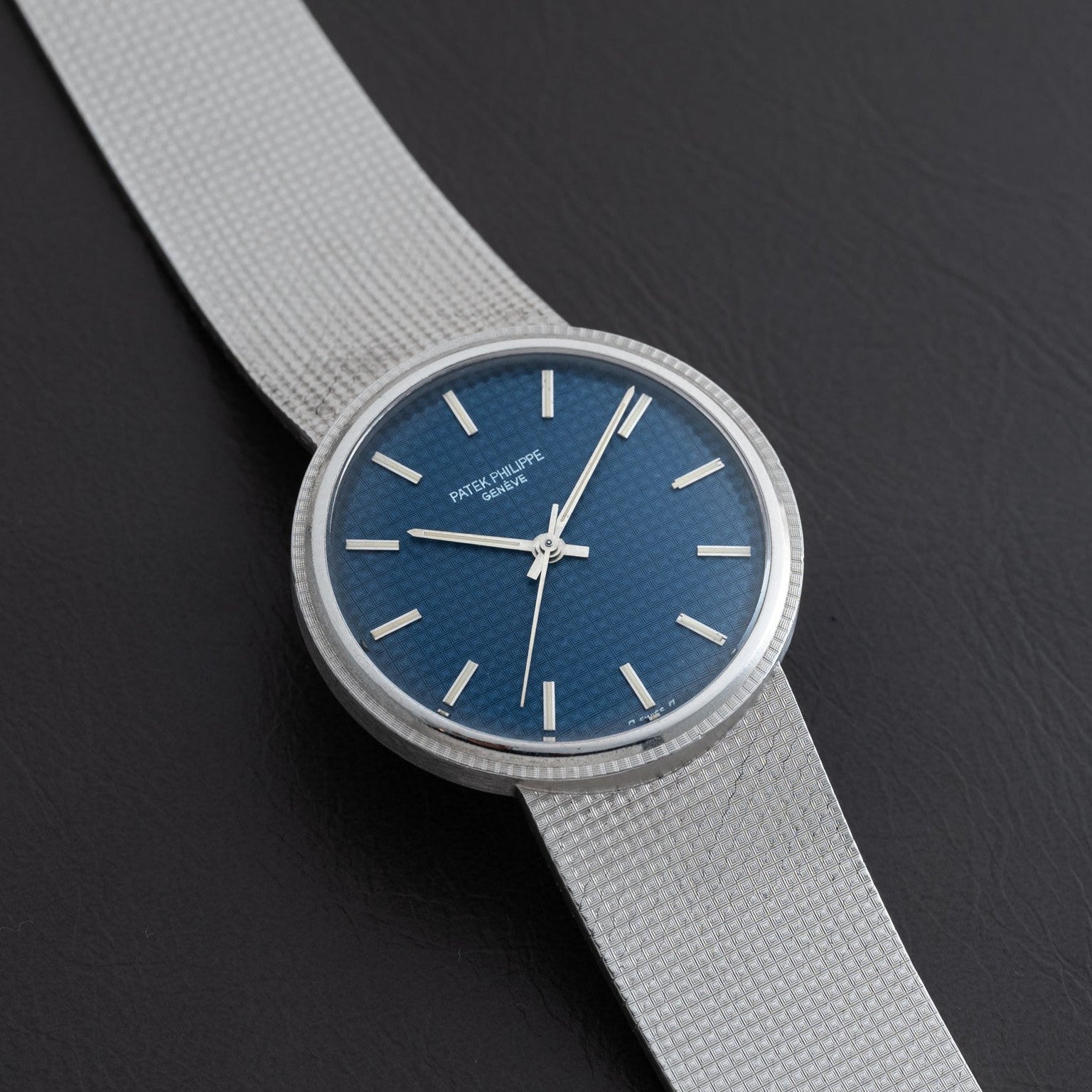 The Patek Philippe Calatrava ‘Backwind’ features a silver case, blue textured face, and silver hour markers, displayed on a dark surface.