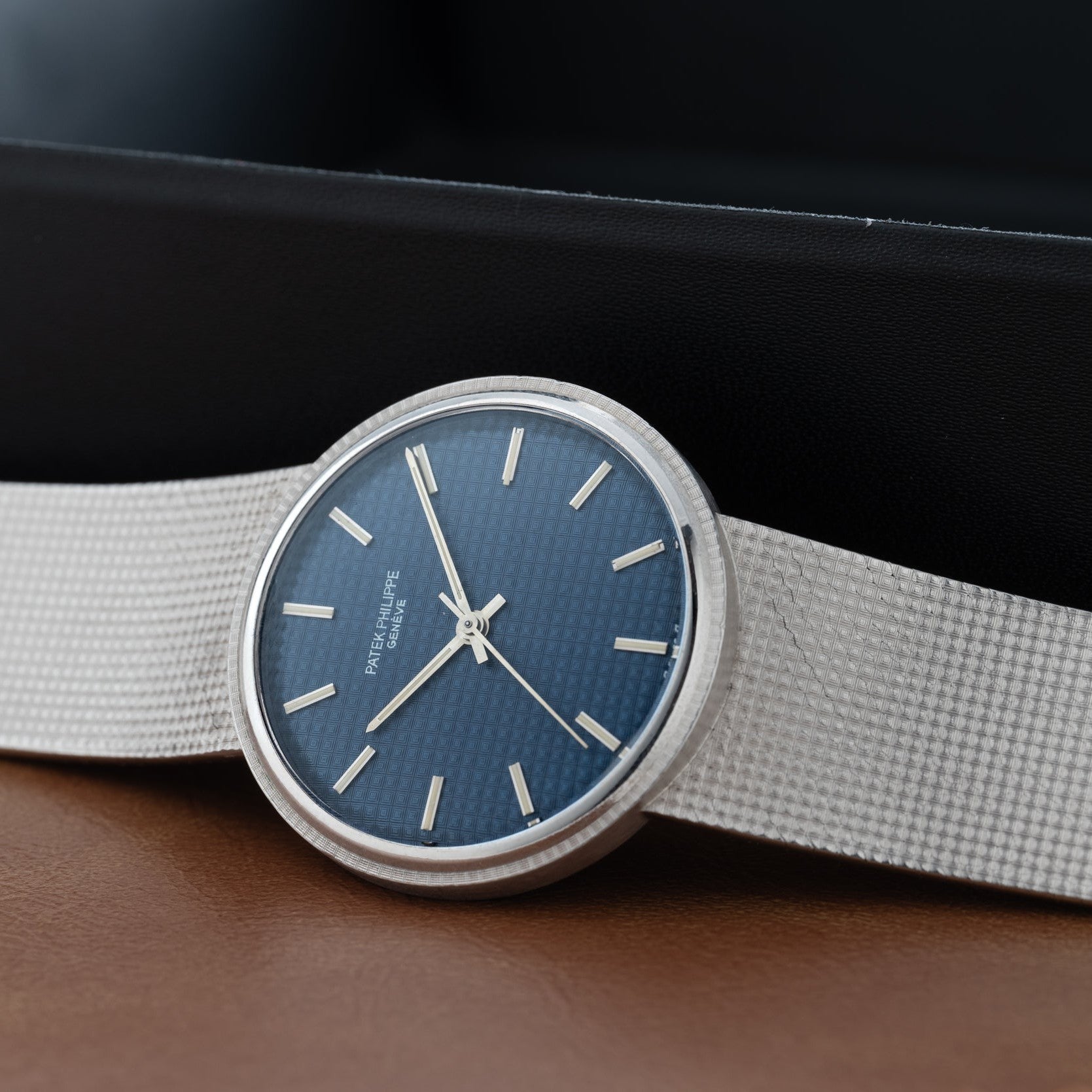 The Patek Philippe Calatrava Backwind features a blue textured dial and white strap, displayed on a brown surface.