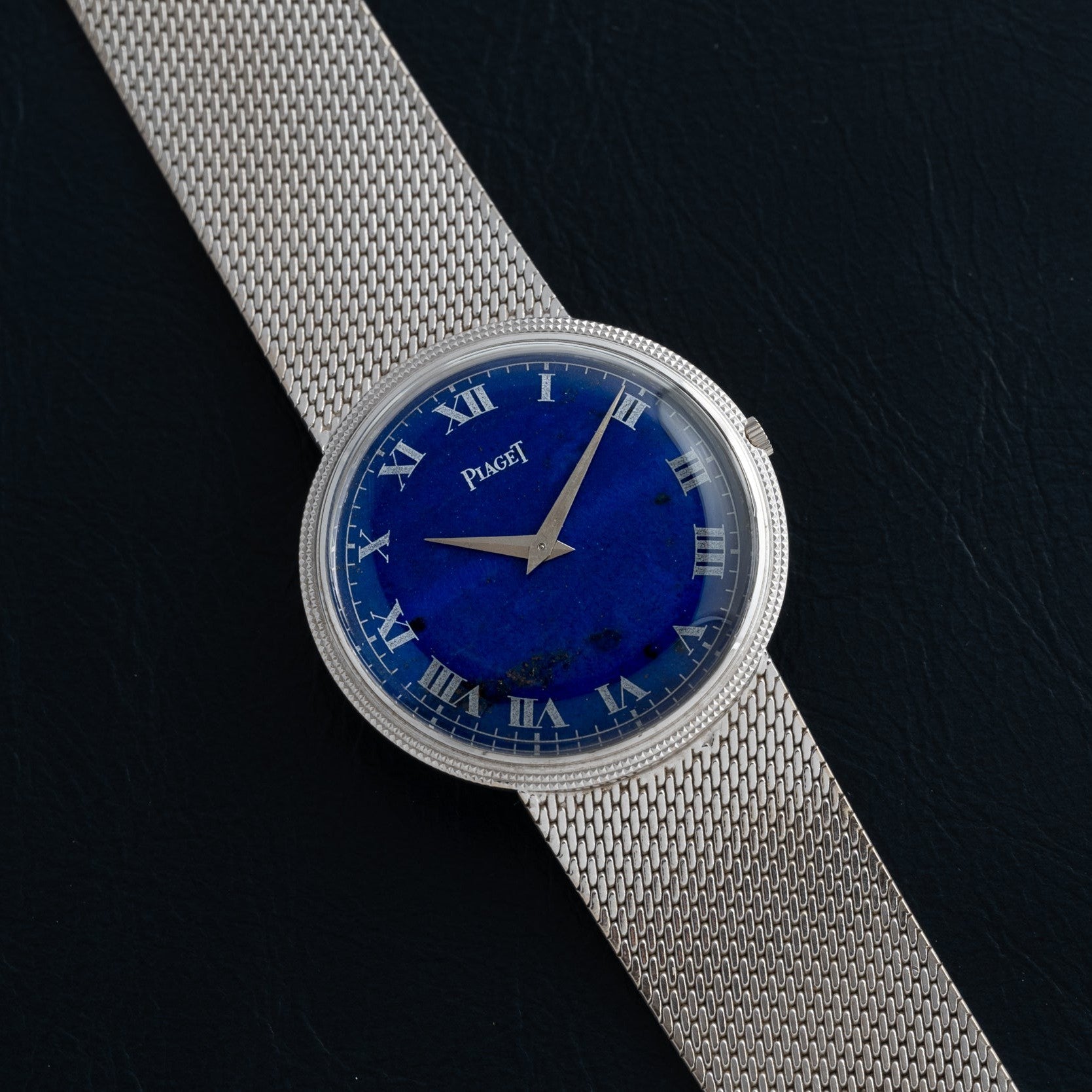 The Piaget Lapis Lazuli Dress Watch features a blue dial with Roman numerals, set in silver and paired with a mesh bracelet, displayed on a black surface.