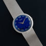 The Piaget Lapis Lazuli Dress Watch features a blue dial with Roman numerals, set in silver and paired with a mesh bracelet, displayed on a black surface.