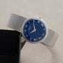 The Piaget Lapis Lazuli Dress Watch features a silver mesh band and blue dial with Roman numerals, shown on a light surface beside a black box.