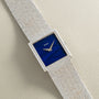 A Piaget White Gold Lapis Lazuli Dress Watch with a rectangular blue face and textured metal band rests on a light textured surface.