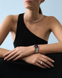 Woman wearing black one-shoulder top with silver watch on wrist and silver neck collar against blue background.
