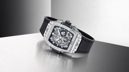 A luxury silver skeleton watch with a black strap, displayed on a reflective metal surface.