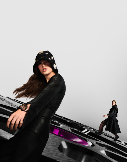 A woman in black with a jeweled hat poses close up; another person walks in the background on a large watch face.