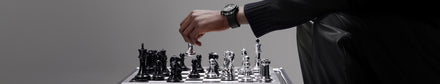 A person’s hand, wearing a watch, moves a chess piece on a metallic chessboard.