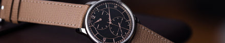 A close-up of a wristwatch with a brown leather strap and a black dial showing rose-gold markings and hands.