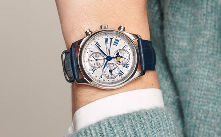 A silver wristwatch with a blue leather strap on a persons wrist, featuring multiple dials and Roman numerals.