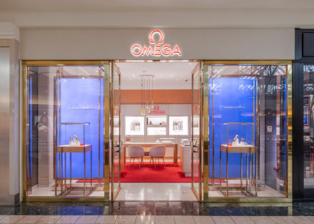 Front view of an Omega store with glass doors, blue displays, and a red carpeted entrance inside a mall.