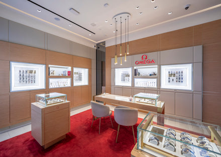Omega watch boutique with display cases, bright lighting, red carpet, and two grey chairs for customers.