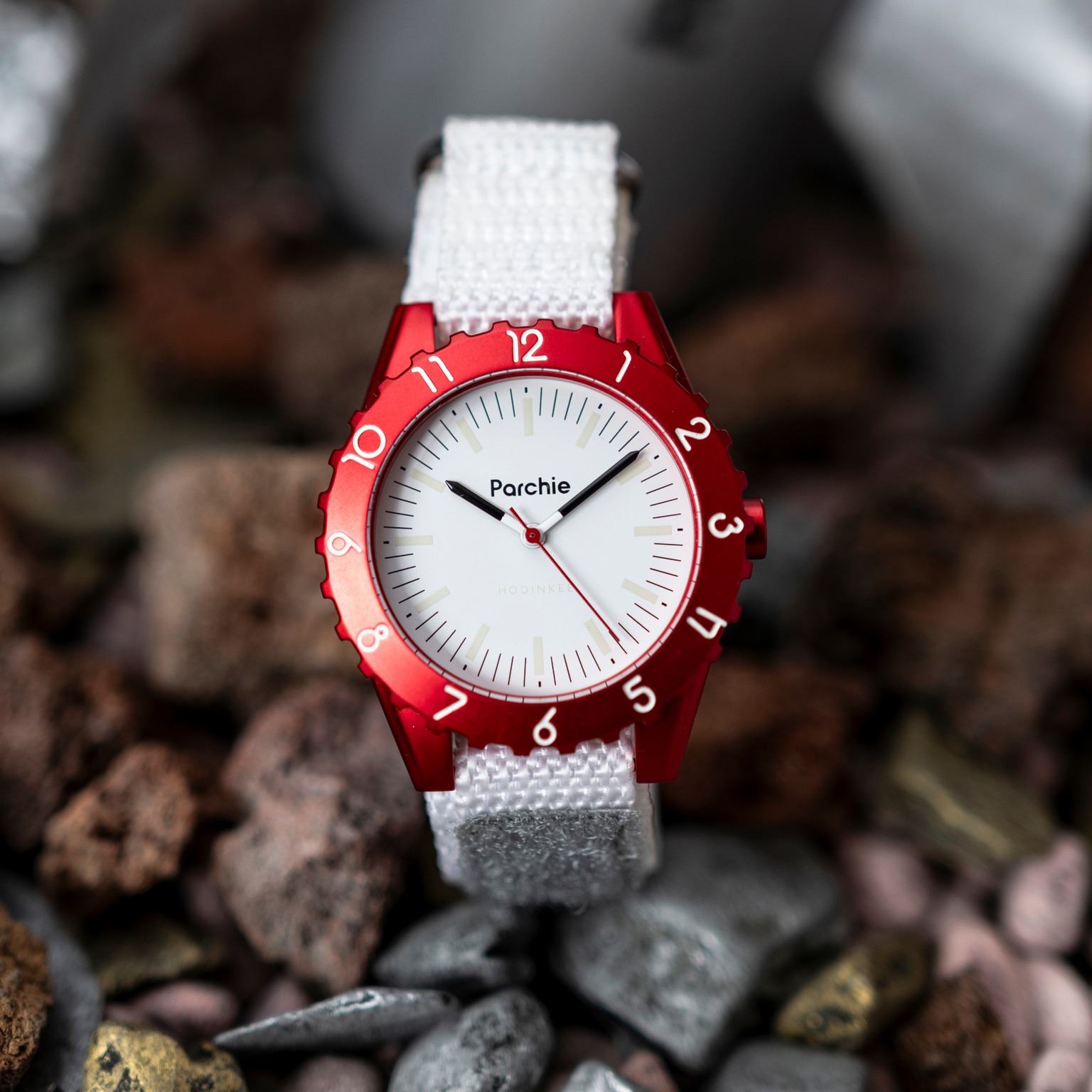 The Parchie Lunar-Time Limited Editions for Hodinkee 32, a white watch with a red bezel and white strap, rests on a surface of mixed rocks and pebbles.