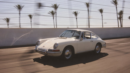 A classic white sports car driving on a sunny road lined with palm trees.