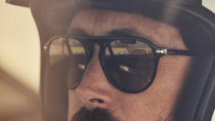 Close-up of a man wearing sunglasses and a helmet, with a racetrack reflected in his lenses.