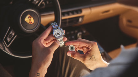 Person holding a luxury wristwatch inside a car with a Porsche logo visible on the steering wheel.