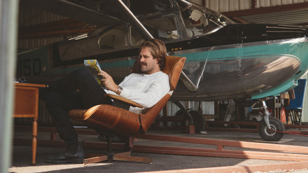 A man sits in a chair reading a map beside a small plane in a hangar.