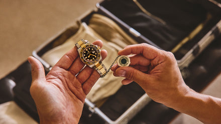 Close-up of hands holding a luxury wristwatch and watch tool above an open suitcase with clothes.