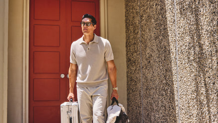 Man in beige polo and pants holding luggage and a jacket, standing by a red door and textured wall.