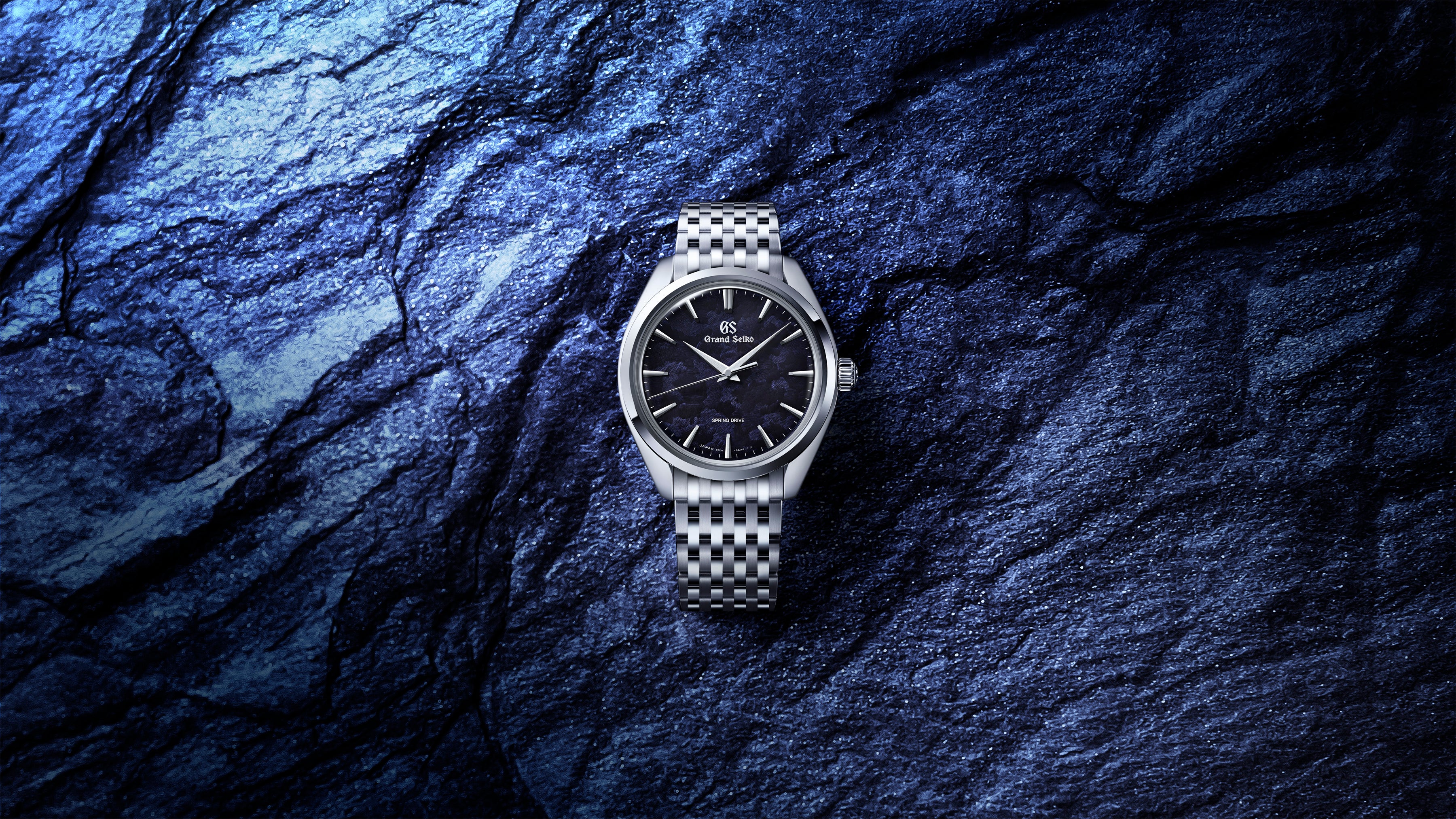 Silver-tone watch with black dial and checkered metal band on blue textured stone background.