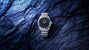 Silver-tone watch with black dial and checkered metal band on blue textured stone background.