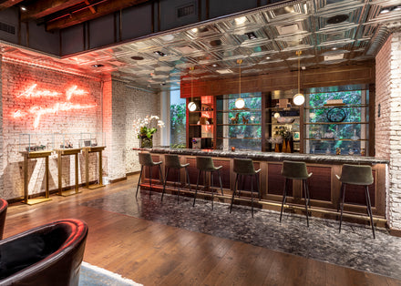 Chic modern bar with velvet stools, brick walls, neon sign, and shelves of bottles behind the counter.
