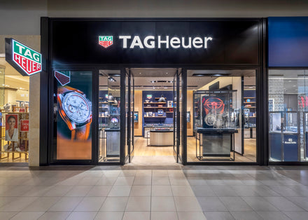 Exterior view of a TAG Heuer store with watches displayed and illuminated brand signage above the entrance.