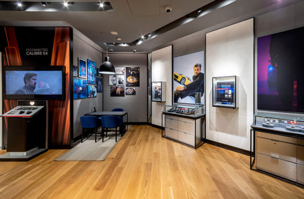 Modern showroom with watches on display, large posters, wooden floor, and a table with blue chairs.