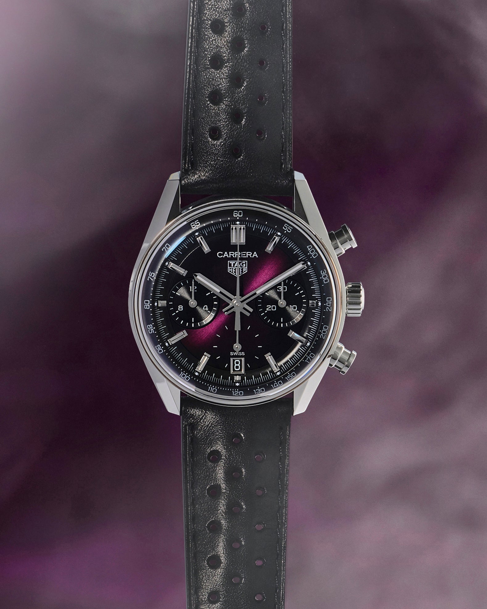 A black TAG Heuer Carrera watch with a perforated leather strap on a purple smoky background.