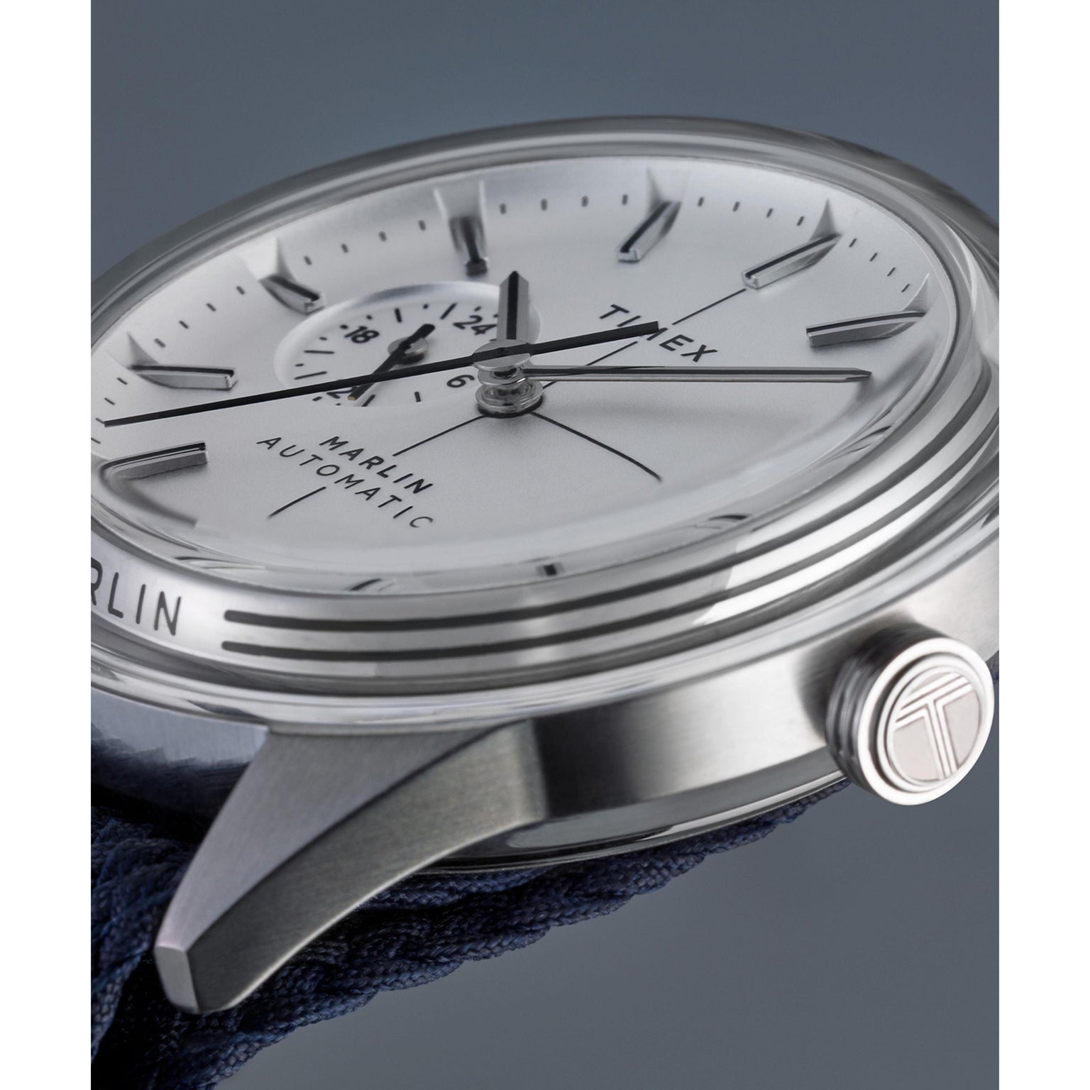 Close-up side view of the Timex Marlin® Jet Automatic watch in silver, featuring a white face, black hour markers, textured crown, and domed Hesalite crystal. Its retro-futuristic design stands out against a bold blue backdrop.