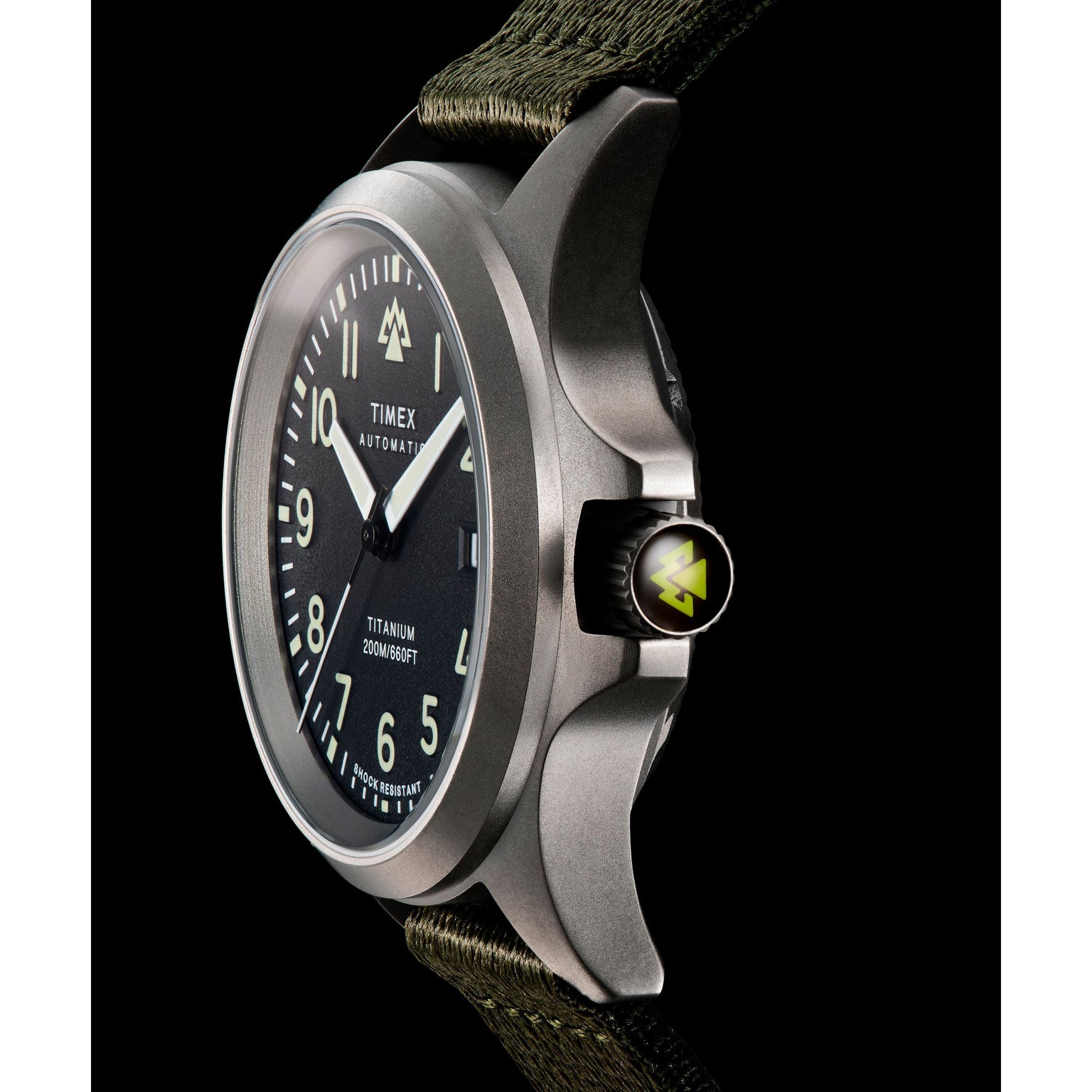A close-up of the Timex Expedition Titanium Automatic watch, featuring a black dial, luminous hands and markers, titanium case, green fabric strap, and a crown with a yellow triangle logo—highlighting its Japanese automatic movement.