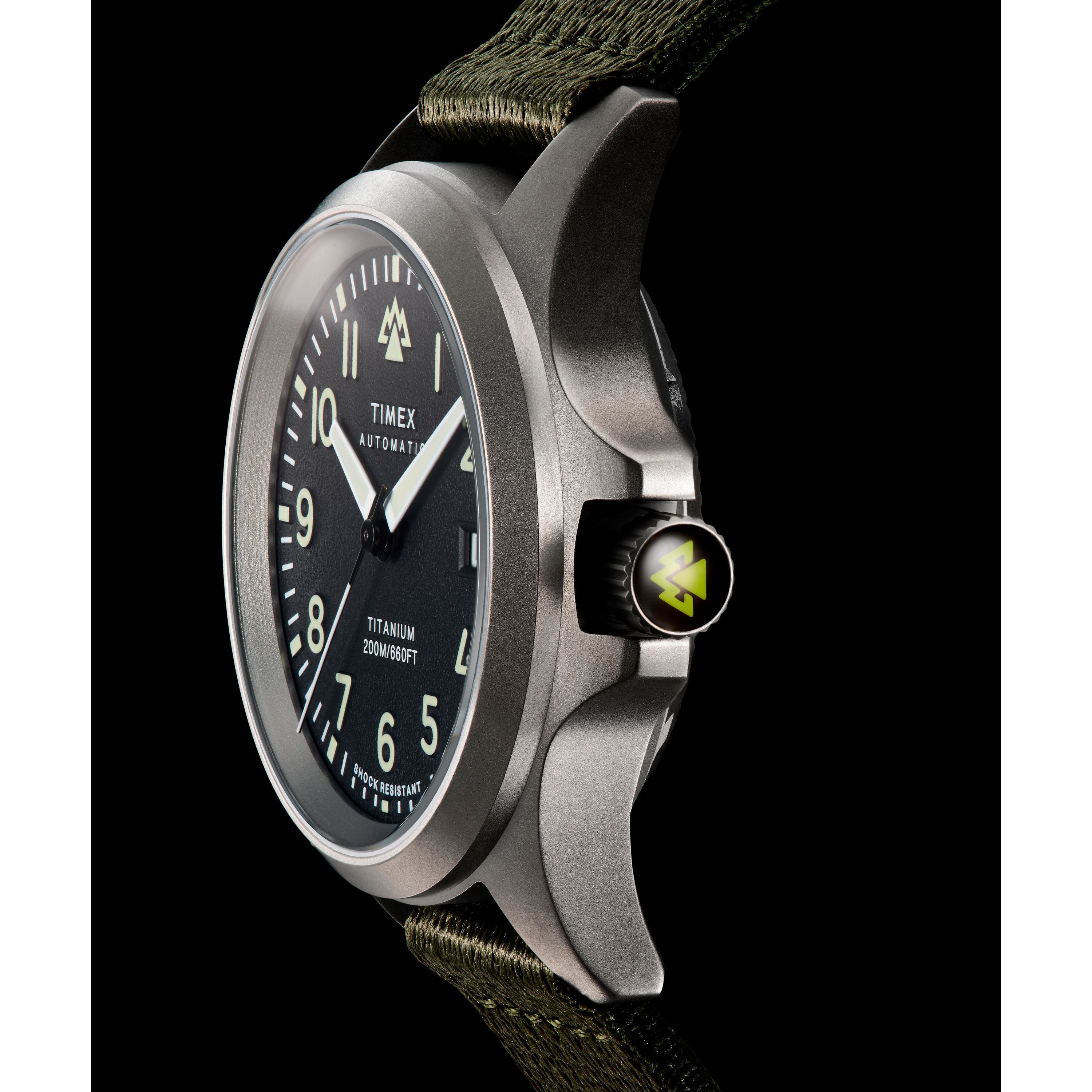 A close-up of the Timex Expedition Titanium Automatic watch, featuring a black dial, luminous hands and markers, titanium case, green fabric strap, and a crown with a yellow triangle logo—highlighting its Japanese automatic movement.