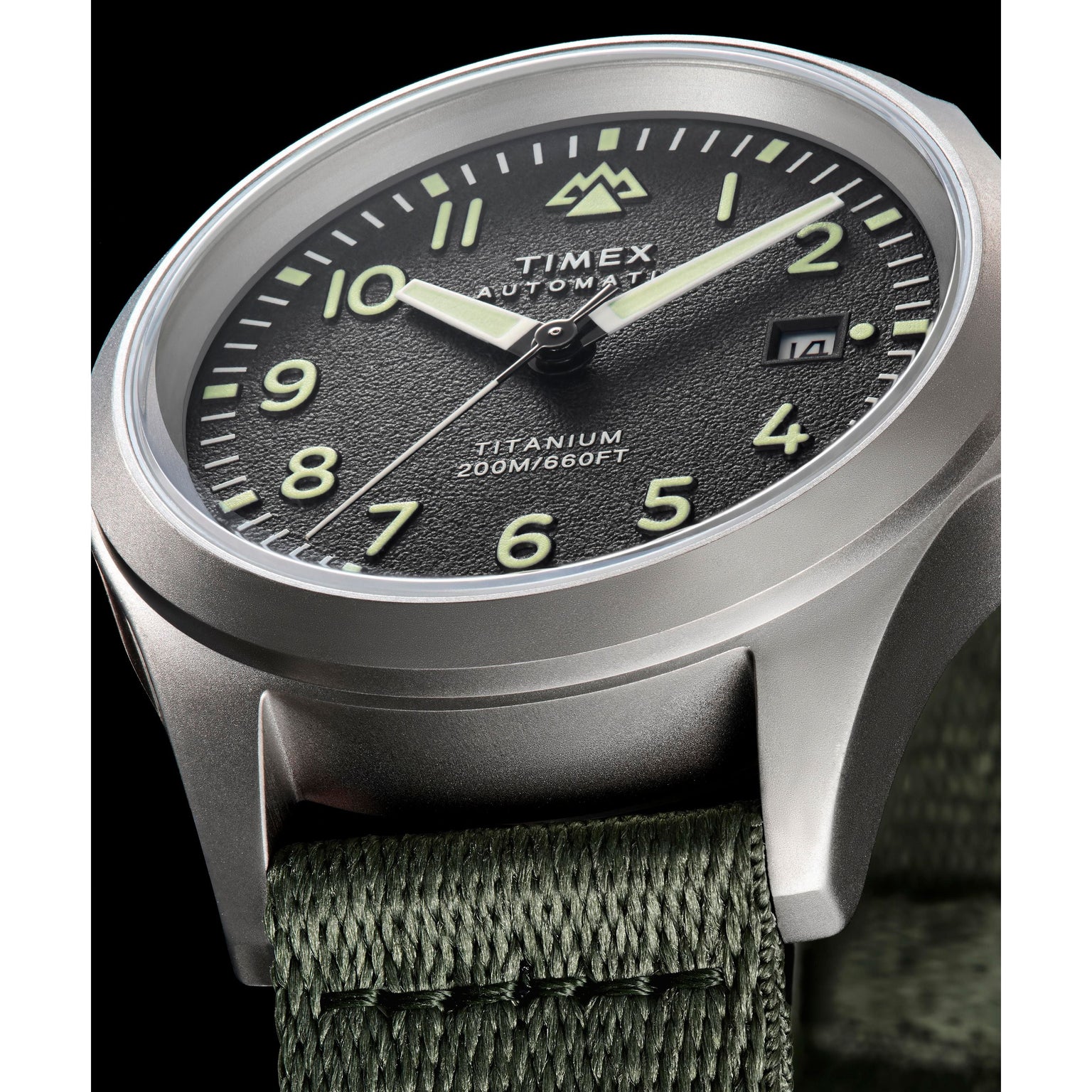 Close-up of the Timex Expedition Titanium Automatic watch with a black textured dial, luminous hands and numerals, date window, titanium 200M/660FT text, Japanese automatic movement, green fabric strap, and silver case.